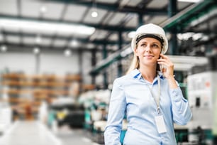 Um retrato de uma jovem engenheira industrial com smartphone e capacete branco em pé em uma fábrica, fazendo um telefonema. Espaço de cópia.