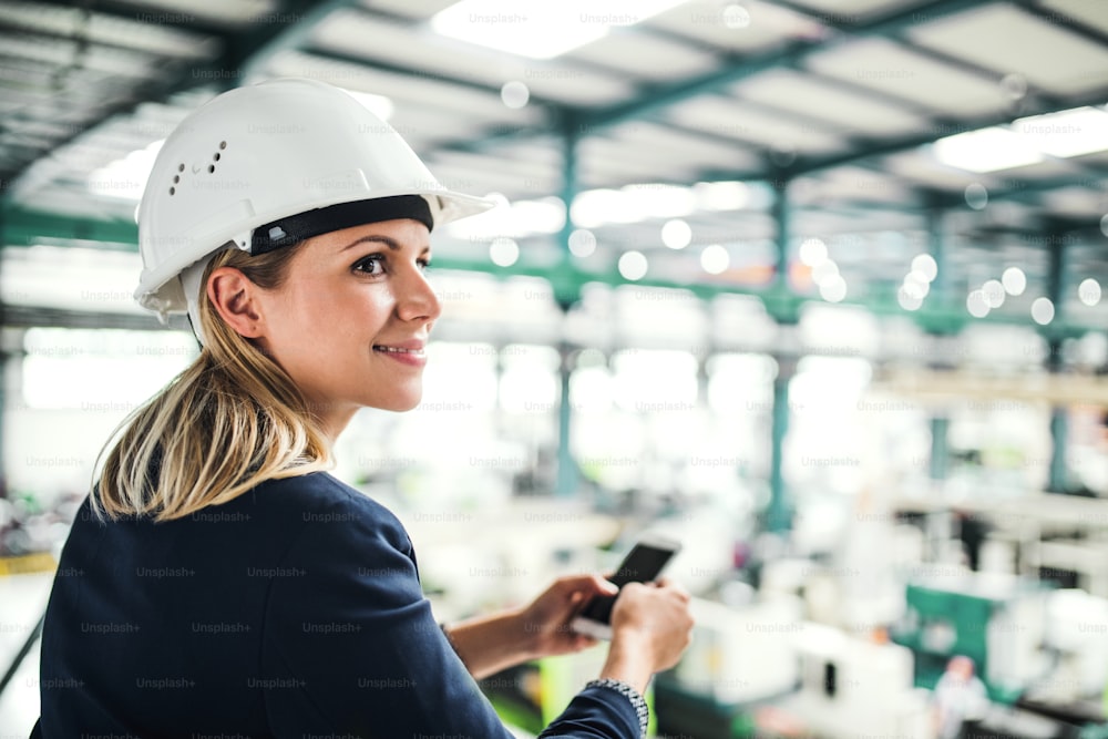Un ritratto di una felice ingegnere industriale in piedi in una fabbrica, con in mano uno smartphone.