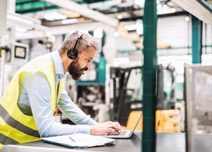 A portrait of a mature industrial man engineer with headset and laptop in a factory, working.
