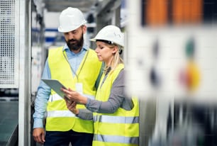 Portrait d’un industriel mûr et d’une ingénieure avec tablette dans une usine, en train de parler.