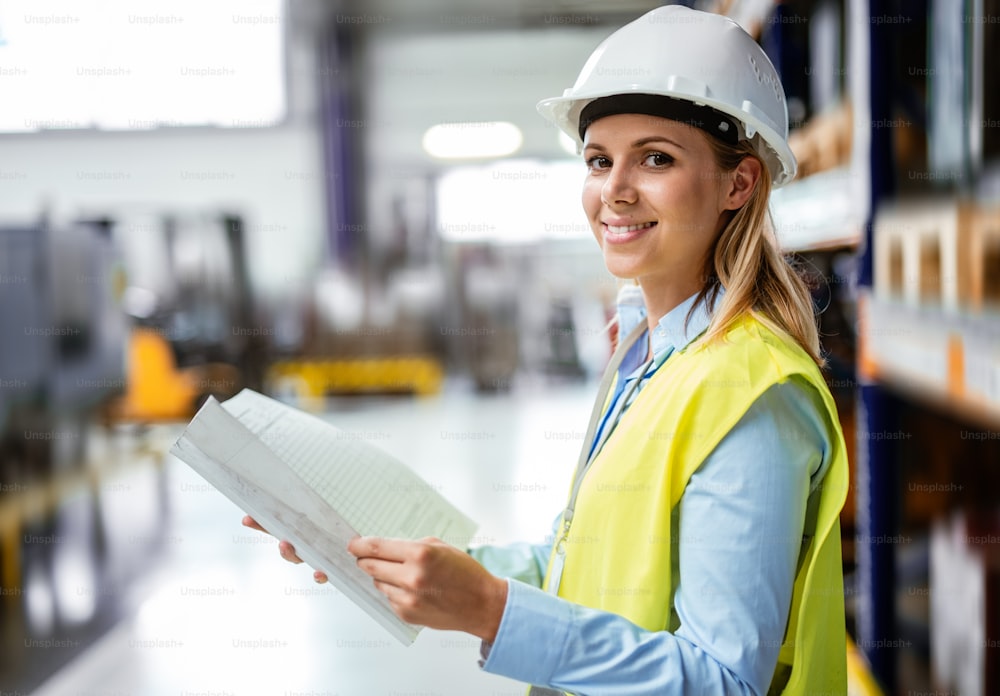 Ein Porträt einer jungen Industrieingenieurin, die in einer Fabrik steht und Blaupausen in der Hand hält.