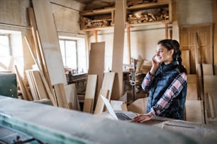 A woman worker with smartphone and laptop in the carpentry workshop, making a phone call.