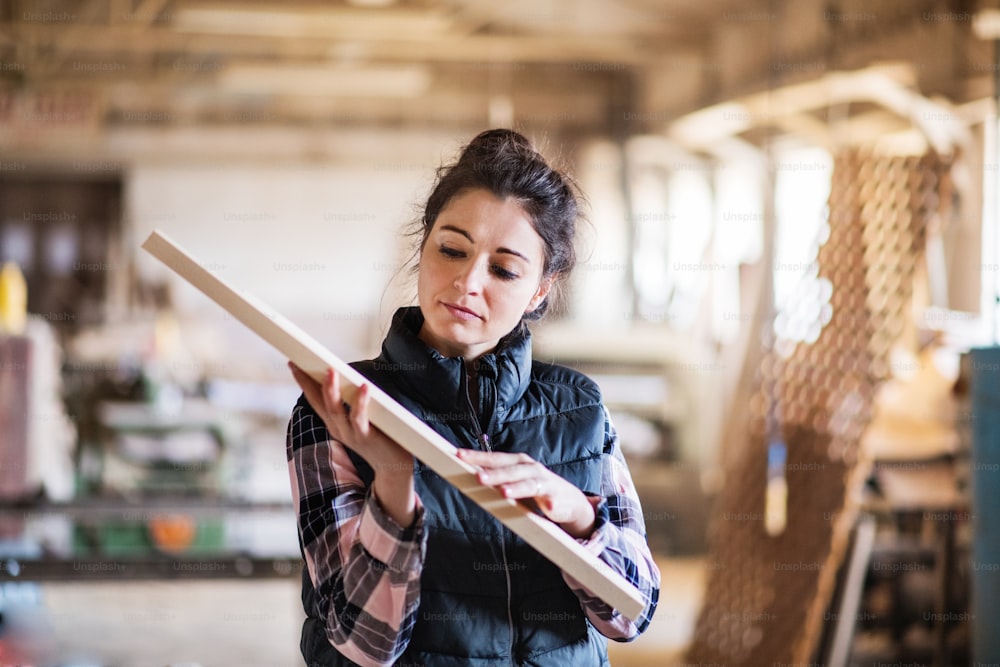 Portrait of a young woman worker in the carpentry workshop, holding a wooden plank.