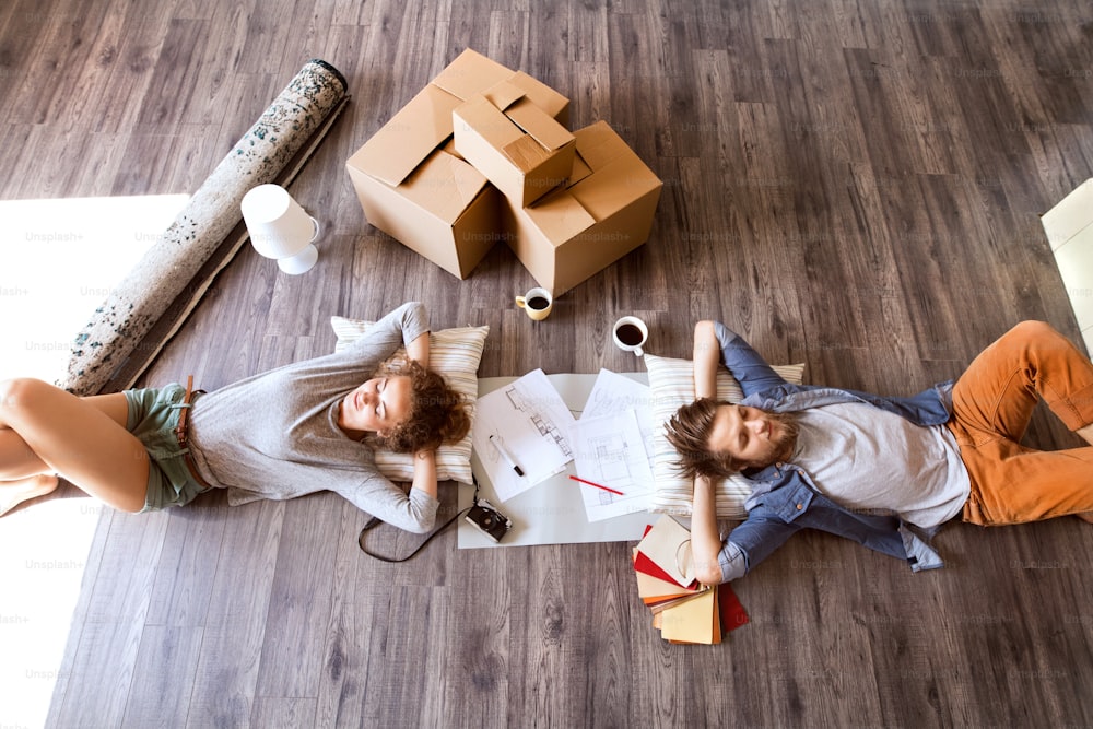 Young married couple moving in new house, lying on the floor near cardboard boxes, drinking coffee.