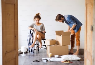 Young married couple moving in a new house, woman with tablet sitting on chair, man unpacking things from a cardboard box.