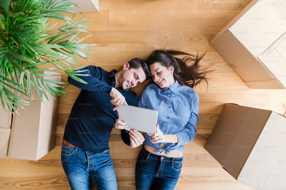 A top view of young couple with tablet and cardboard boxes lying on the floor, moving in a new home.
