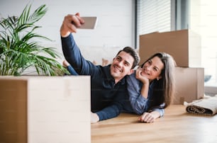 A young happy couple with a smartphone and cardboard boxes lying on a floor, taking selfie when moving in a new home.