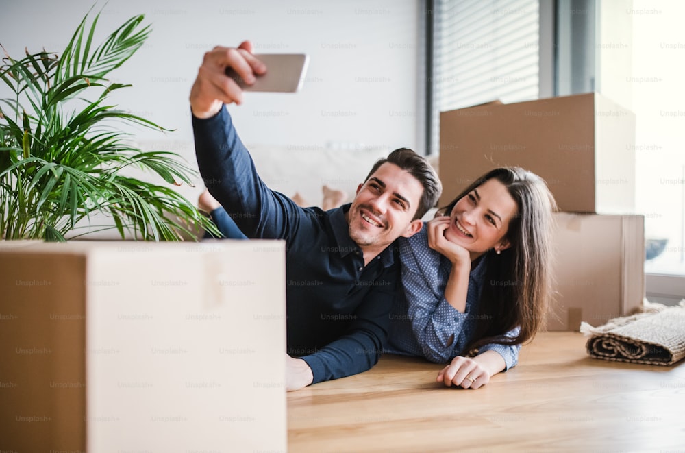 Ein junges glückliches Paar mit einem Smartphone und Pappkartons, das auf dem Boden liegt und ein Selfie macht, wenn es in ein neues Zuhause zieht.