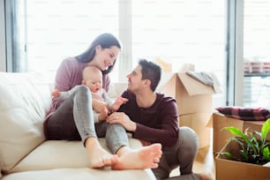 Ein Porträt eines glücklichen jungen Paares mit Baby und Pappkartons, das in ein neues Zuhause zieht.