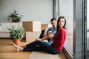 Young happy couple with cardboard boxes moving in a new home, sitting on a floor with coffee.