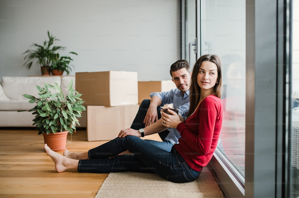Jovem casal feliz com caixas de papelão se movendo em uma nova casa, sentado em um chão com café.