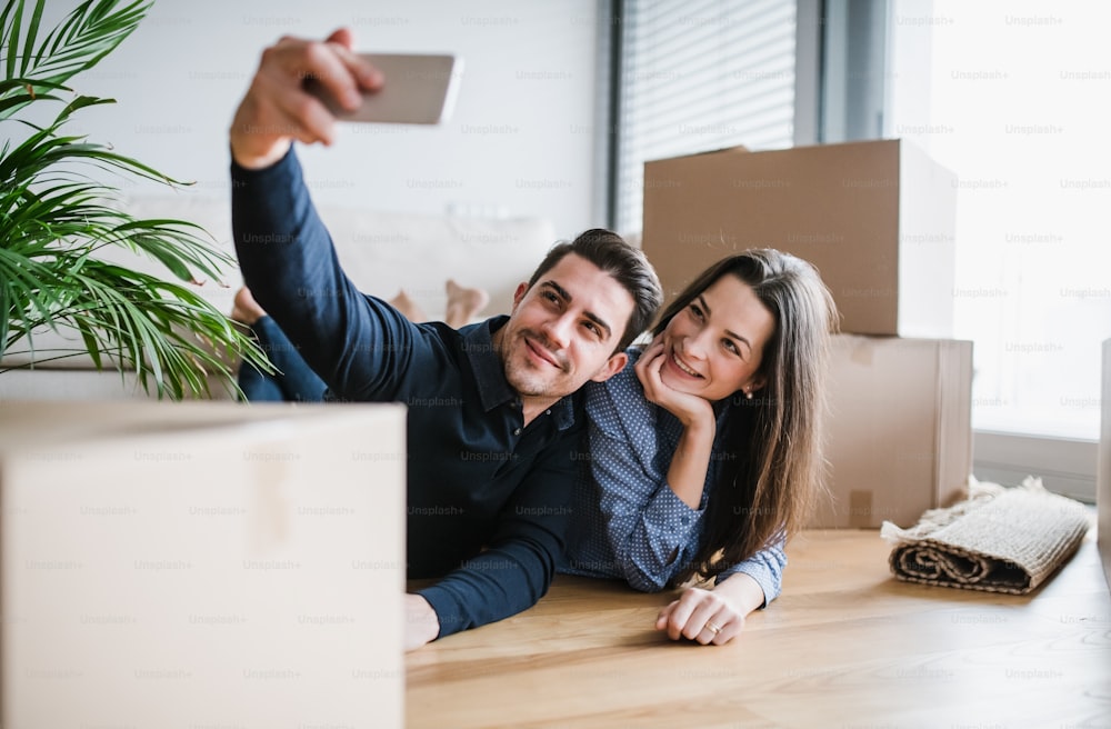 Un jeune couple heureux avec un smartphone et des boîtes en carton allongés sur un sol, prenant un selfie lors de l’emménagement dans une nouvelle maison.