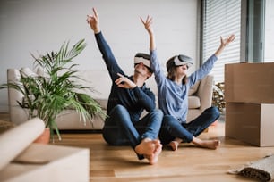 A young couple with VR goggles and cardboard boxes sitting barefoot on a floor, moving in a new home.