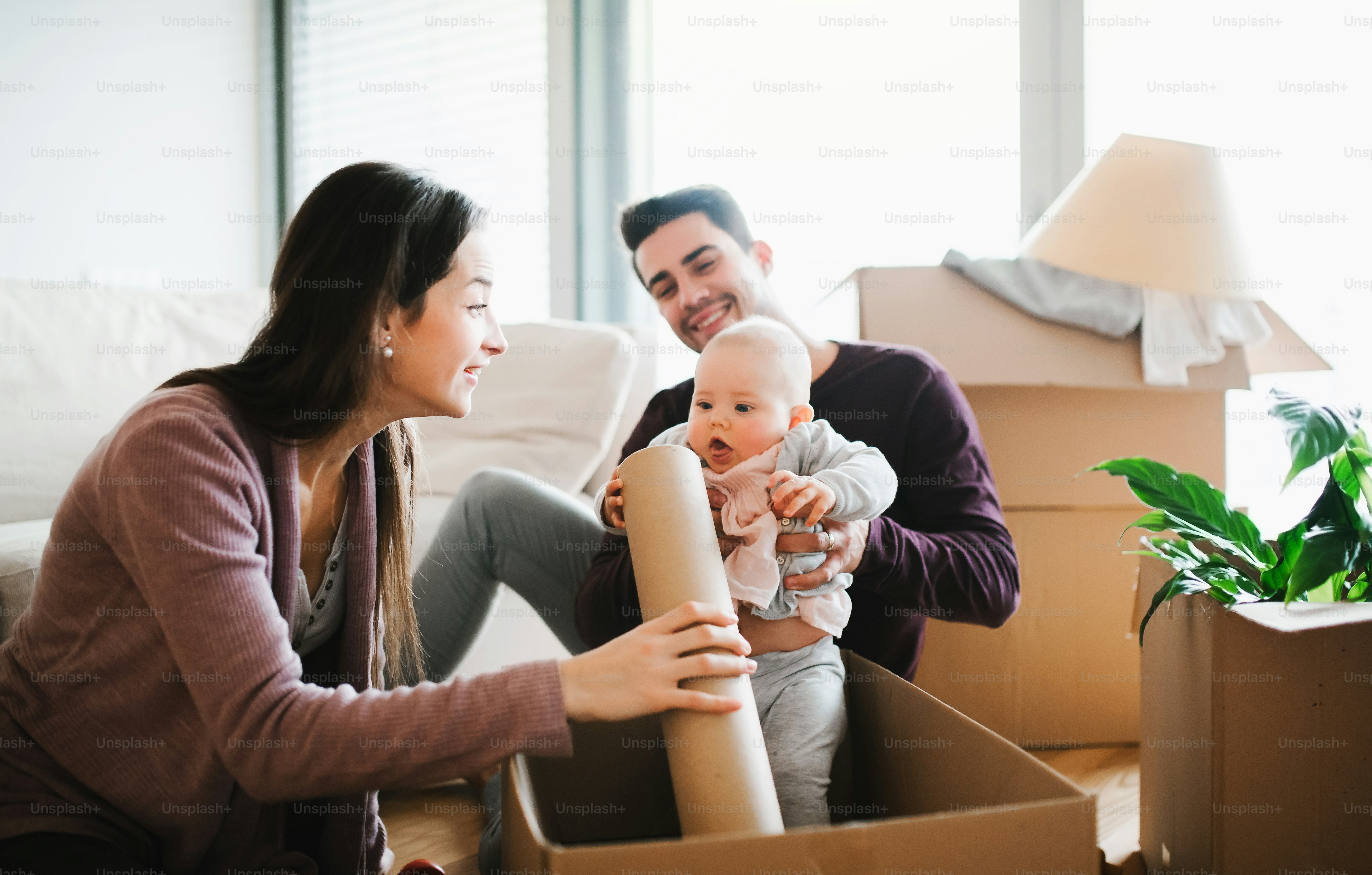 A portrait of happy young couple sitting on a sofa with a baby and cardboard boxes, moving in a new home.
