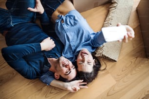 Uma vista superior de um jovem casal feliz com um smartphone e caixas de papelão deitado no chão, tirando selfie ao se mudar para uma nova casa.