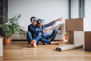 Ein junges Paar mit VR-Brille und Pappkartons sitzt barfuß auf dem Boden und zieht in ein neues Zuhause.