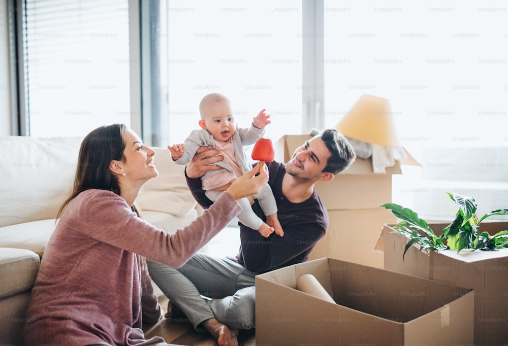 A portrait of happy young couple sitting on the floor with a baby and cardboard boxes, moving in a new home.