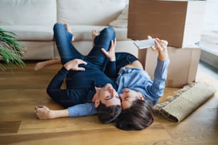 Una joven pareja feliz con un teléfono inteligente y cajas de cartón tirada en el suelo, tomándose selfies cuando se muda a un nuevo hogar.