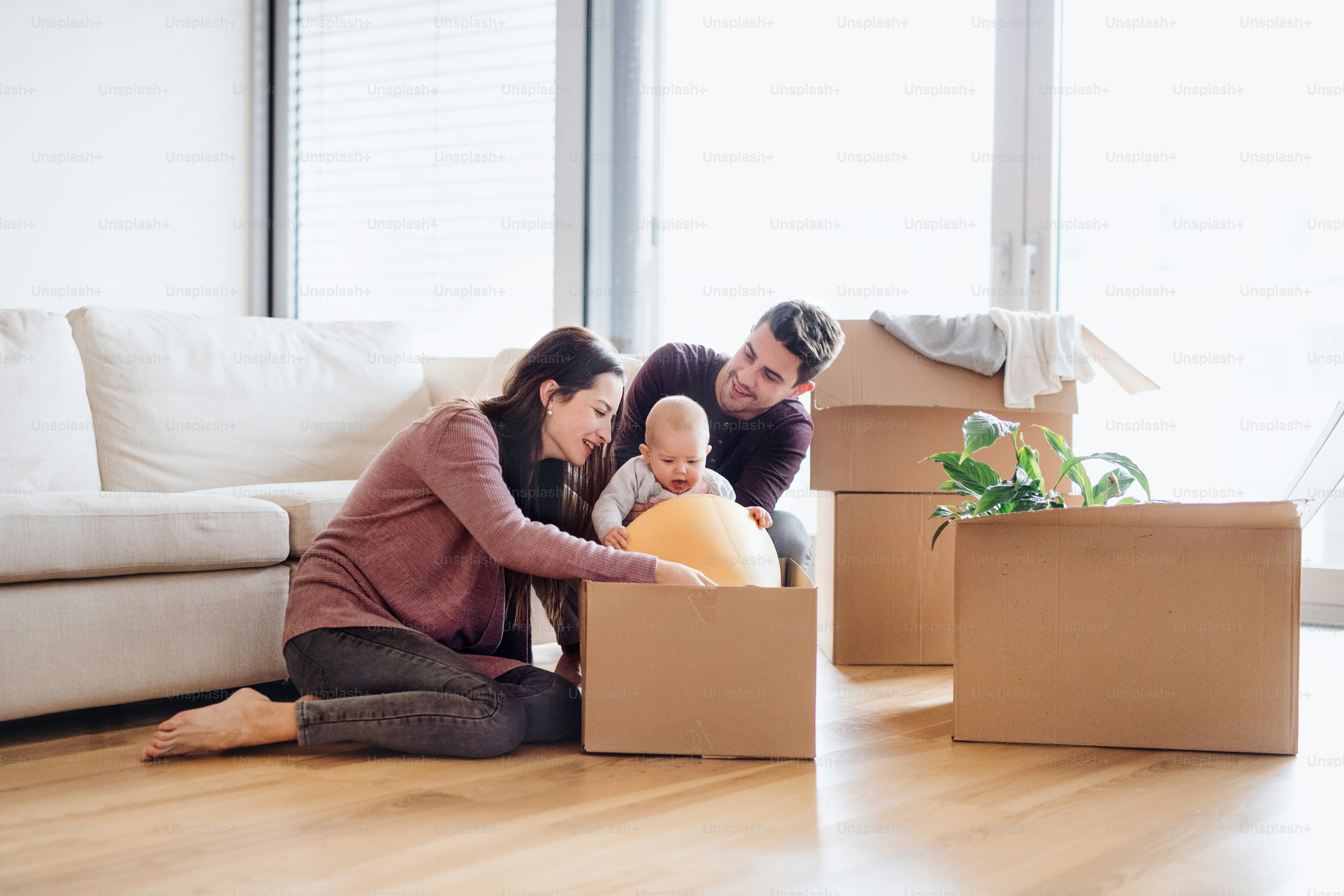 A portrait of happy young couple sitting on a sofa with a baby and cardboard boxes, moving in a new home.