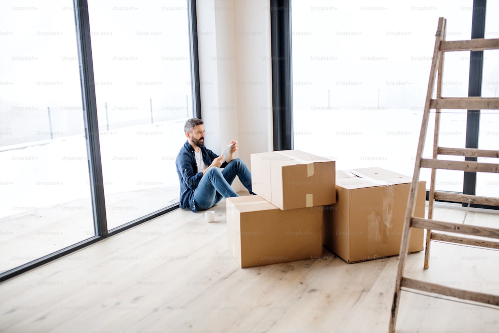 Um homem maduro com caixas de tablet e papelão sentado no chão, mobiliando nova casa. Um novo conceito de casa. Espaço de cópia.