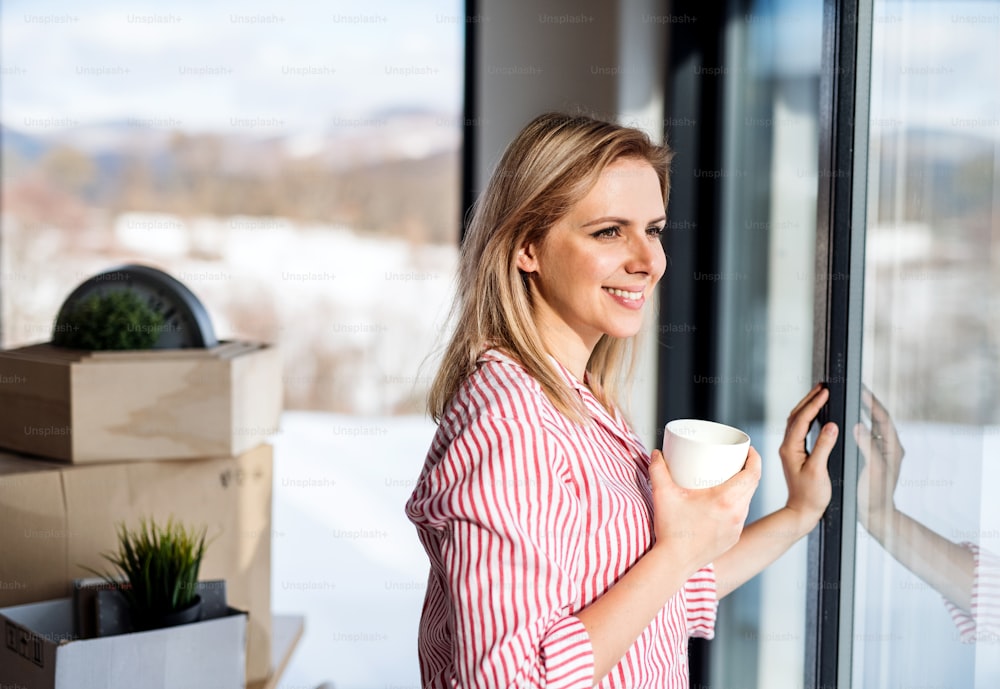 Una giovane donna felice che si trasferisce in una nuova casa, appoggiata a una finestra e bevendo caffè.