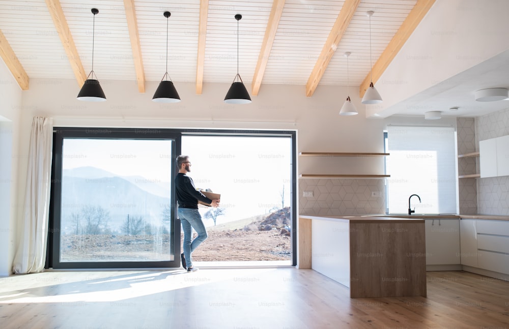 Mature man with moving box standing by patio door in unfurnished house, looking out.