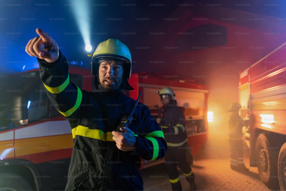 A low angle view of m firefighter talking to walkie talkie with fire truck in background at night.