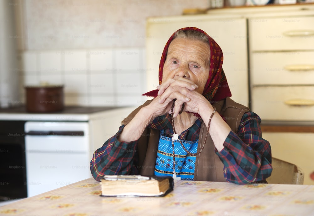 Una mujer muy anciana con pañuelo en la cabeza está rezando en su cocina de estilo campestre