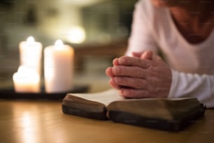 Unrecognizable senior woman lying on the floor praying with hands clasped together on her Bible. Burning candles next to her. Close up.