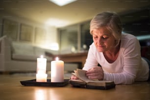 Schöne ältere Frau zu Hause in ihrem Wohnzimmer, auf dem Boden liegend und ihre Bibel lesend. Brennende Kerzen neben ihr. Aufschließen.