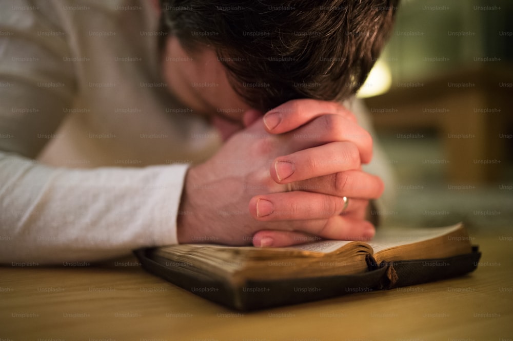 Jeune homme méconnaissable priant, agenouillé sur le sol, les mains sur sa Bible. Gros plan.
