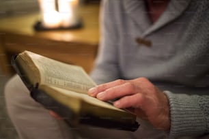 Nicht wiederzuerkennender älterer Mann zu Hause in seinem Wohnzimmer, auf dem Boden sitzend, Bibel lesend. Brennende Kerzen hinter ihm.