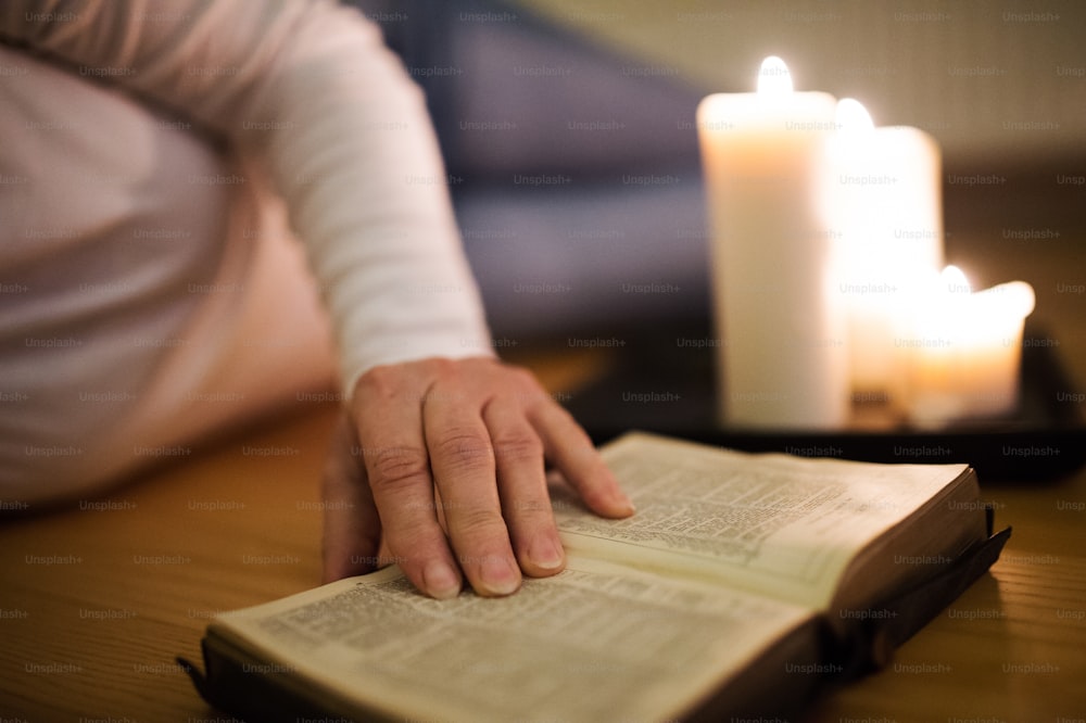 Mujer irreconocible tirada en el suelo leyendo su Biblia. Velas encendidas junto a ella. Primer plano del libro y su mano.