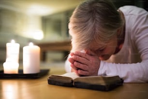 Mujer mayor arrodillada en el suelo orando con las manos juntas sobre su Biblia. Velas encendidas junto a ella. Cerrar.