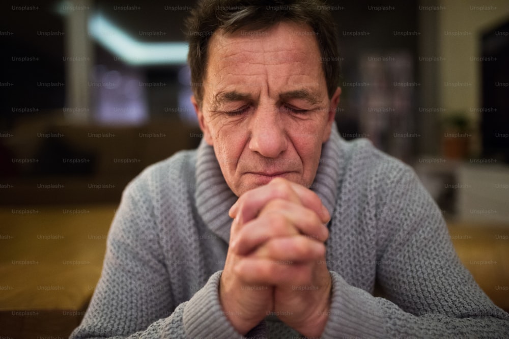 Hombre mayor con suéter gris en casa en su sala de estar rezando, con las manos juntas, los ojos cerrados