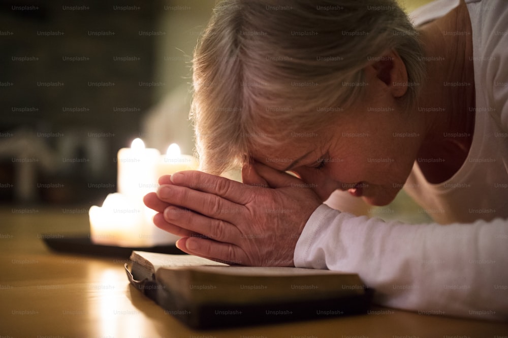 Femme âgée agenouillée sur le sol en train de prier, les mains jointes sur sa Bible. Des bougies allumées à côté d’elle. Gros plan.