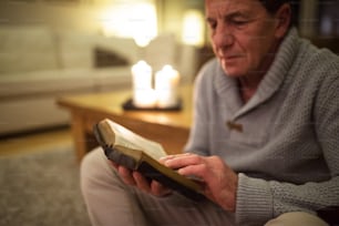 Älterer Mann zu Hause in seinem Wohnzimmer, auf dem Boden sitzend, Bibel lesend. Brennende Kerzen hinter ihm.