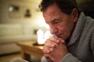 Senior man in gray sweater at home in his living room praying, hands clasped together, eyes closed. Burning candles behind him. Close up.