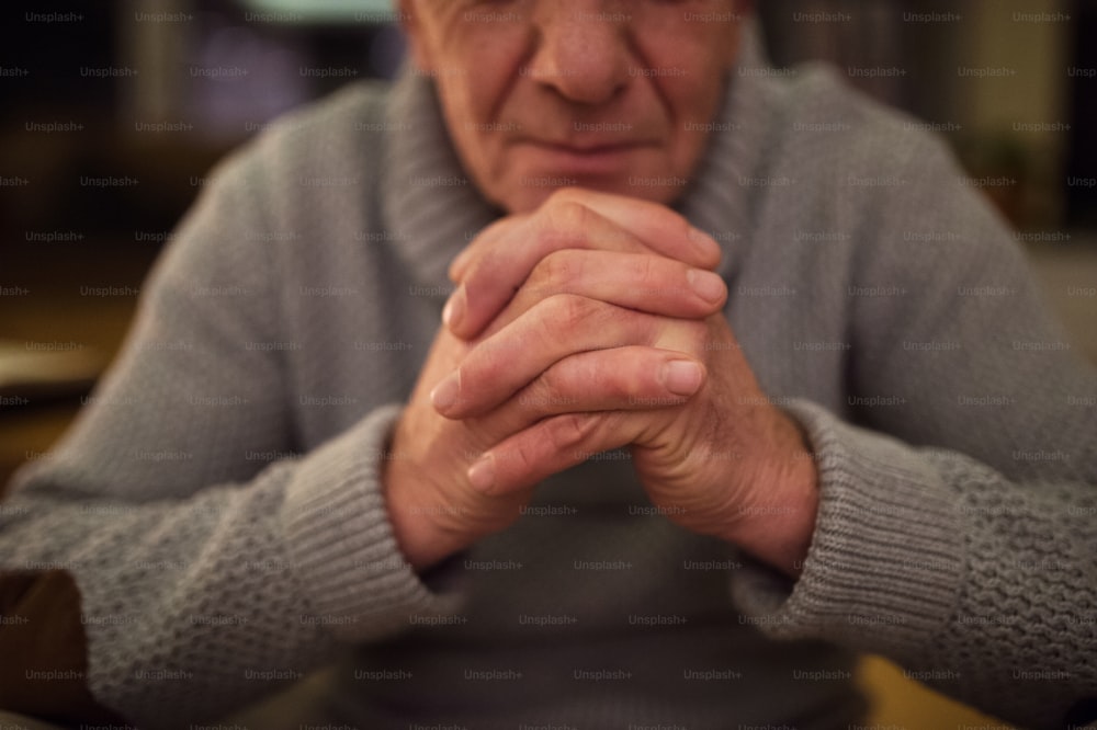 Uomo anziano irriconoscibile in maglione grigio a casa nel suo salotto che prega, mani giunte, occhi chiusi