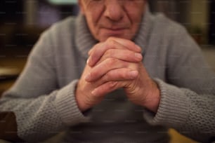 Homem idoso irreconhecível de camisola cinzenta em casa na sua sala de estar a rezar, mãos entrelaçadas, olhos fechados