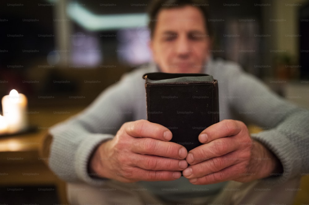 Senior man praying, holding an old Bible in his hands. Burning candles behind him. Close up.