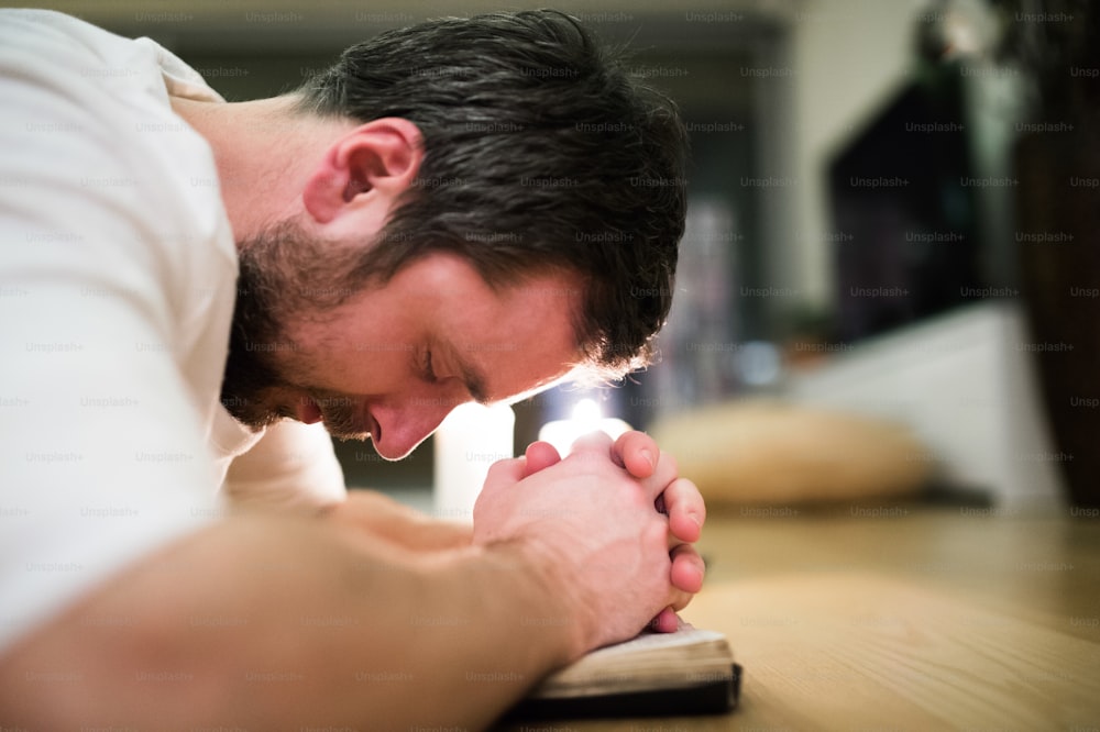 Hübscher junger Mann, der betet, auf dem Boden kniet und seine Bibel in den Händen hält. Brennende Kerzen hinter ihm. Aufschließen.