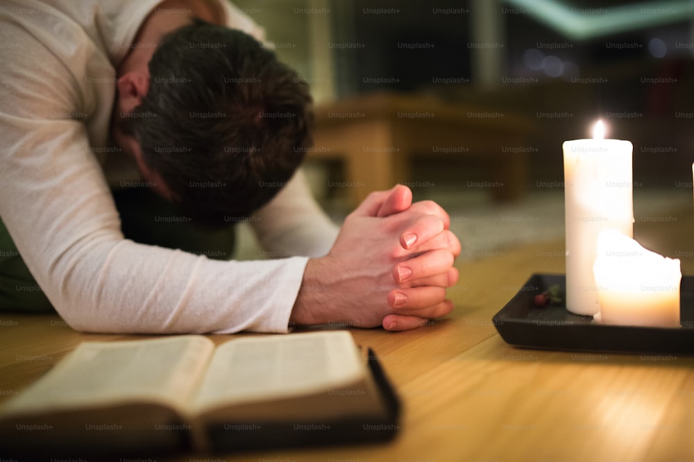 Jeune homme méconnaissable en train de prier, agenouillé sur le sol, les mains jointes. Bible et bougies allumées à côté de lui. Gros plan.