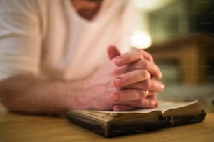 Unrecognizable young man praying, kneeling on the floor, hands on his Bible. Close up.