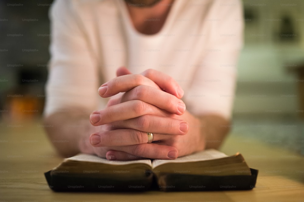 Unrecognizable young man praying, kneeling on the floor, hands on his Bible. Close up.