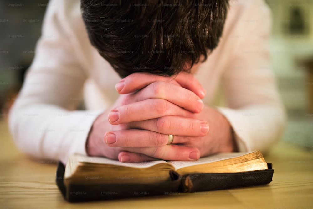 Unrecognizable young man praying, kneeling on the floor, hands on his Bible. Close up.