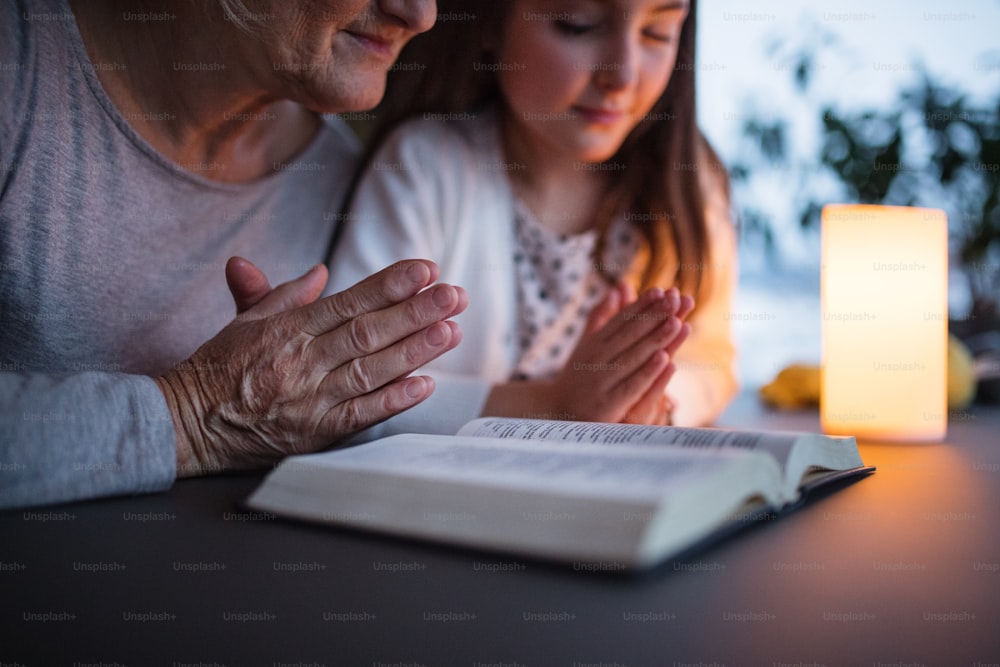 Una bambina e la sua irriconoscibile nonna con la Bibbia a casa, pregando. Concetto di famiglia e generazioni.