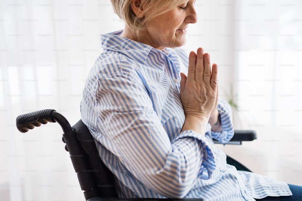 Una mujer mayor en silla de ruedas rezando en casa, con los ojos cerrados.