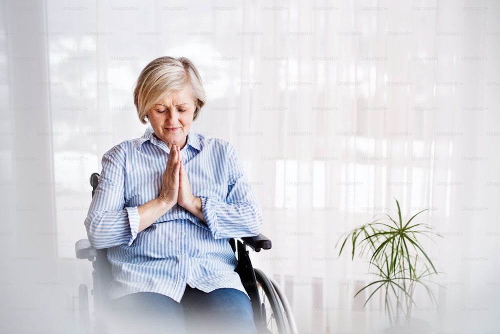 Una mujer mayor en silla de ruedas rezando en casa, con los ojos cerrados.
