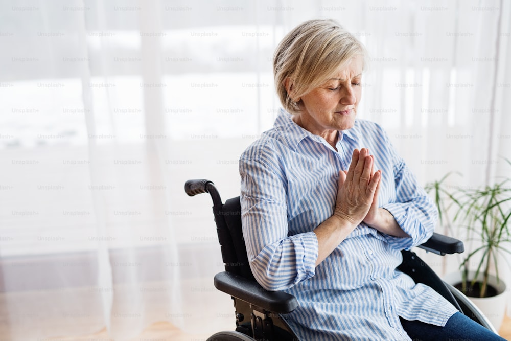 Eine ältere Frau im Rollstuhl betet zu Hause mit geschlossenen Augen.